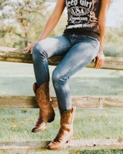 female sitting on a woodenfence with cowboybots and jeans with a black t-shirt with white writing infront. The image is from upper torsov and down