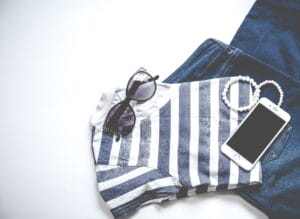 T-shirt with sunglases laying on top of a pair of jeans with a bracelet and phone